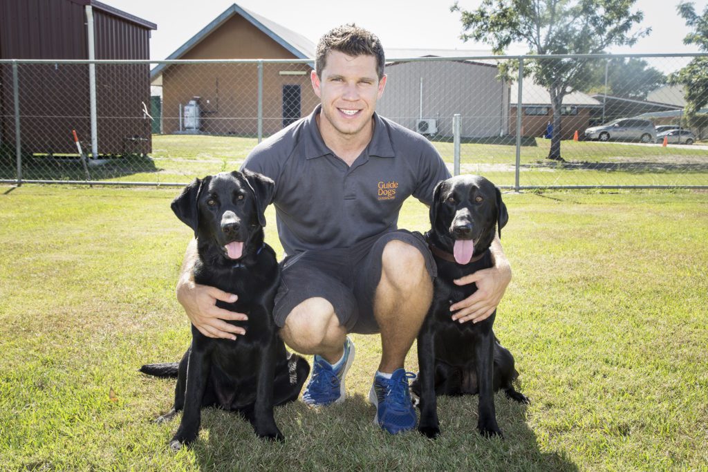 Image of a male Guide Dog Trainer crouching between two black Guide Dogs