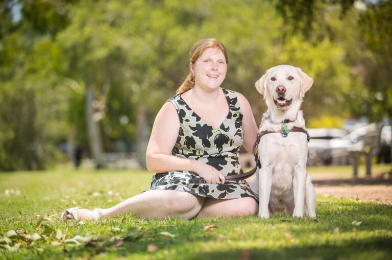 Image of a young woman sitting beside her yellow Guide Dog on the grass