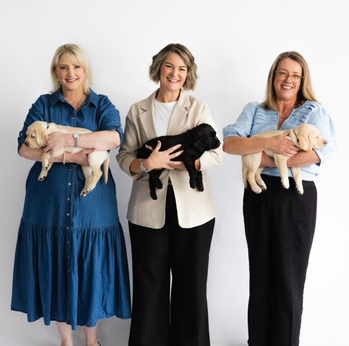 Image shows Corinne (Left), Renae (Middle) and Liz (Right) holding Labrador puppies and smiling to the camera.