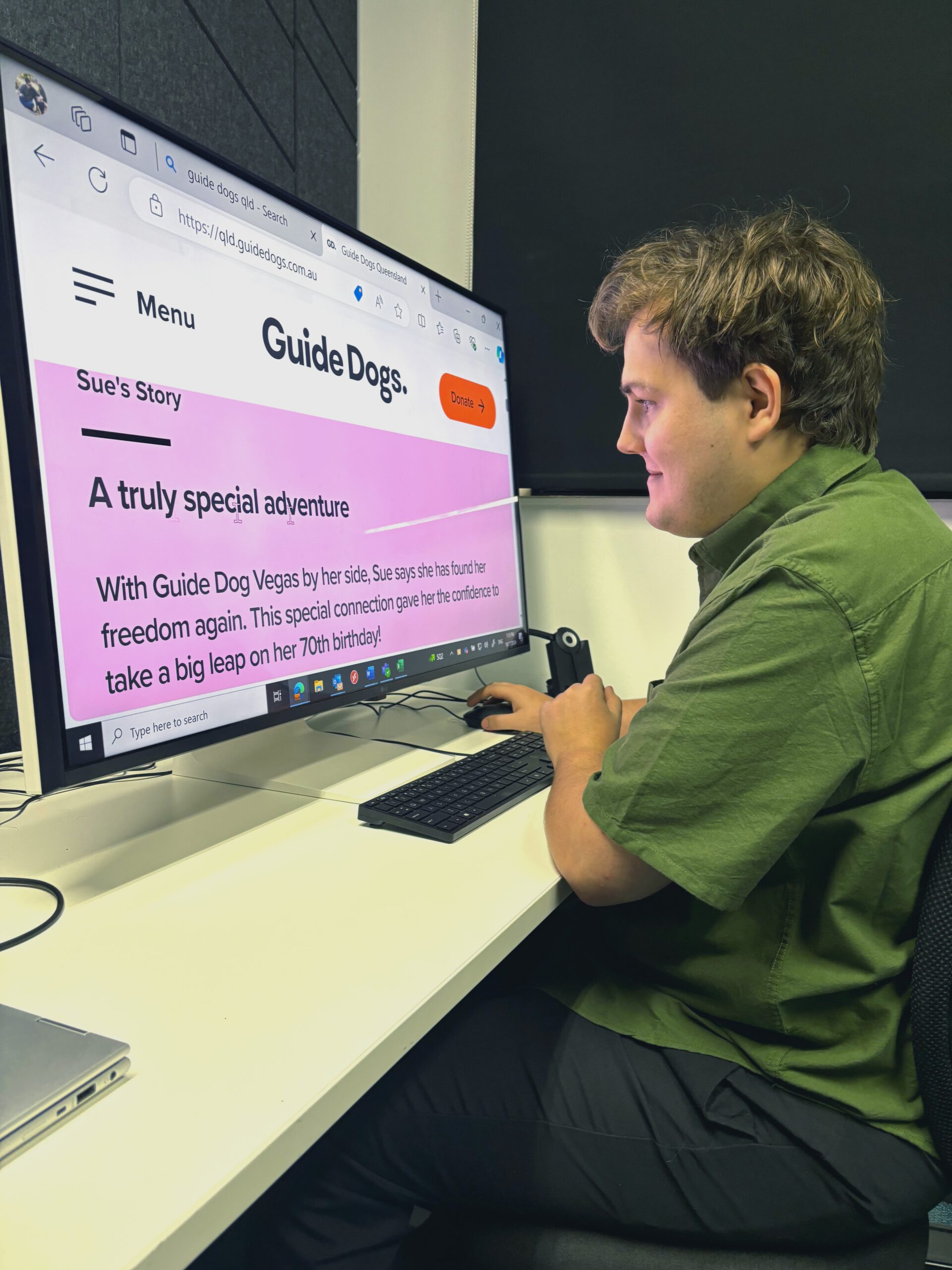 Image shows Brent, seated at a desk using a large computer screen with enlarged text displaying a Guide Dogs website. The screen shows a heading 