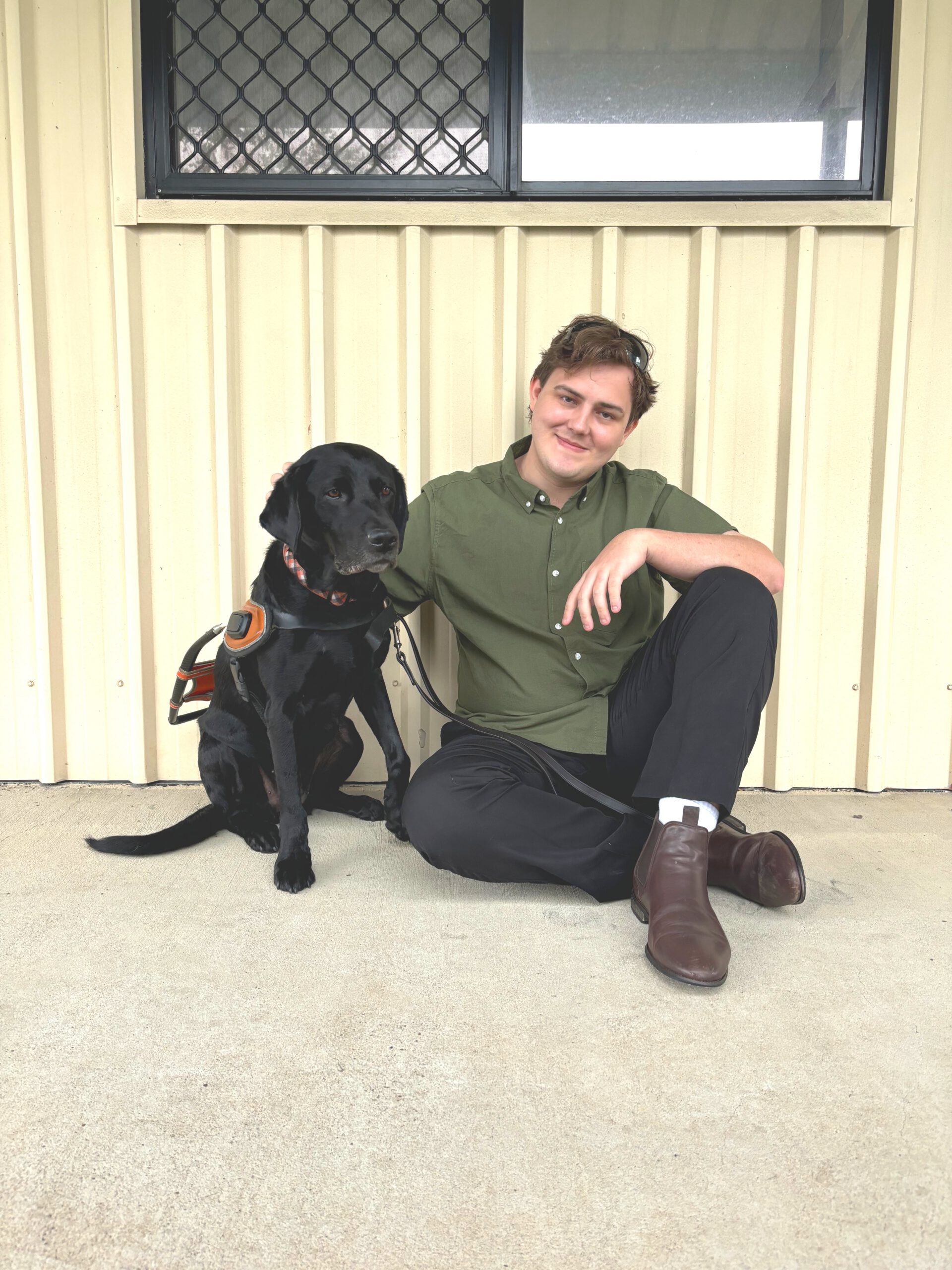 Image shows Brent smiling at the camera while sitting on the ground next to his Guide Dog, Jaycee, who is wearing a guide harness. They are in front of a beige wall with a window above.