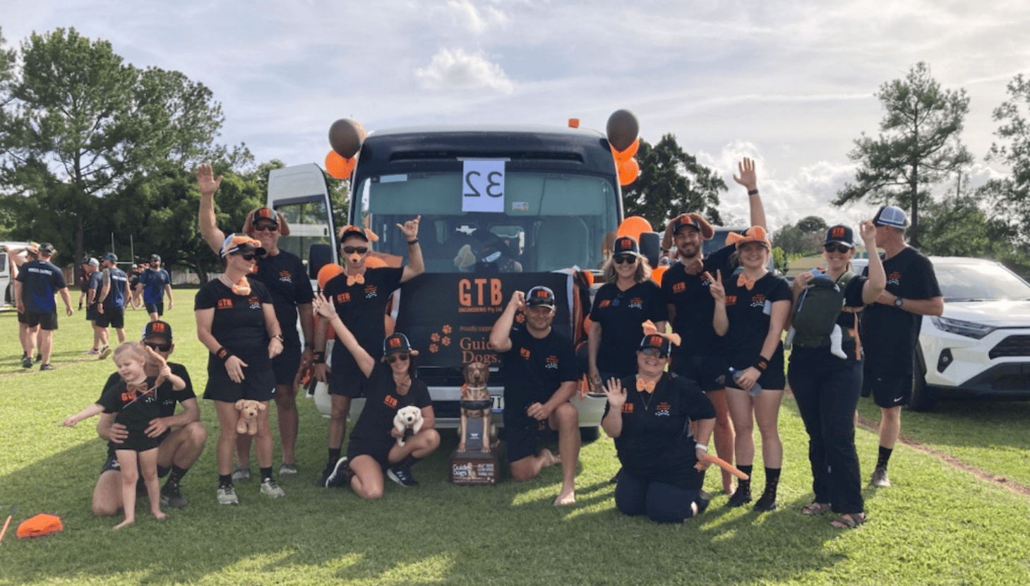 Image shows a group of people in matching black GTB shirts posing in front of a decorated bus with orange and brown balloons, holding up peace signs and guide dog plush toys at a community event.