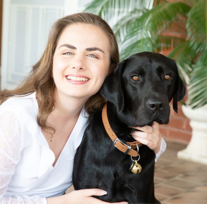 A person looking at the camera smiling with their arm around the neck of a black labrador dog.