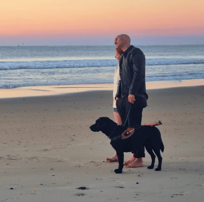 Guide dogs client; Rhett standing on the beach during a sunset, with his black Guide Dog next to him.