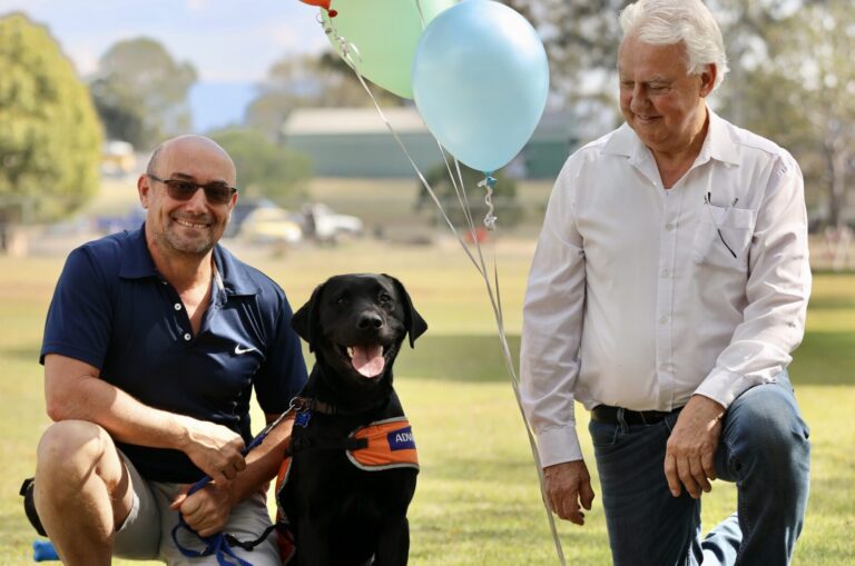 John from Baron Barnett Lodge kneeling alongside a bunch of orange, green, blue and silver birthday balloons as well as Guide Dog-in-training, Baron, and his Puppy Raiser.