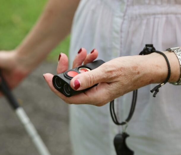 Person standing outside with a white cane in one hand and a Miniguide in the other.