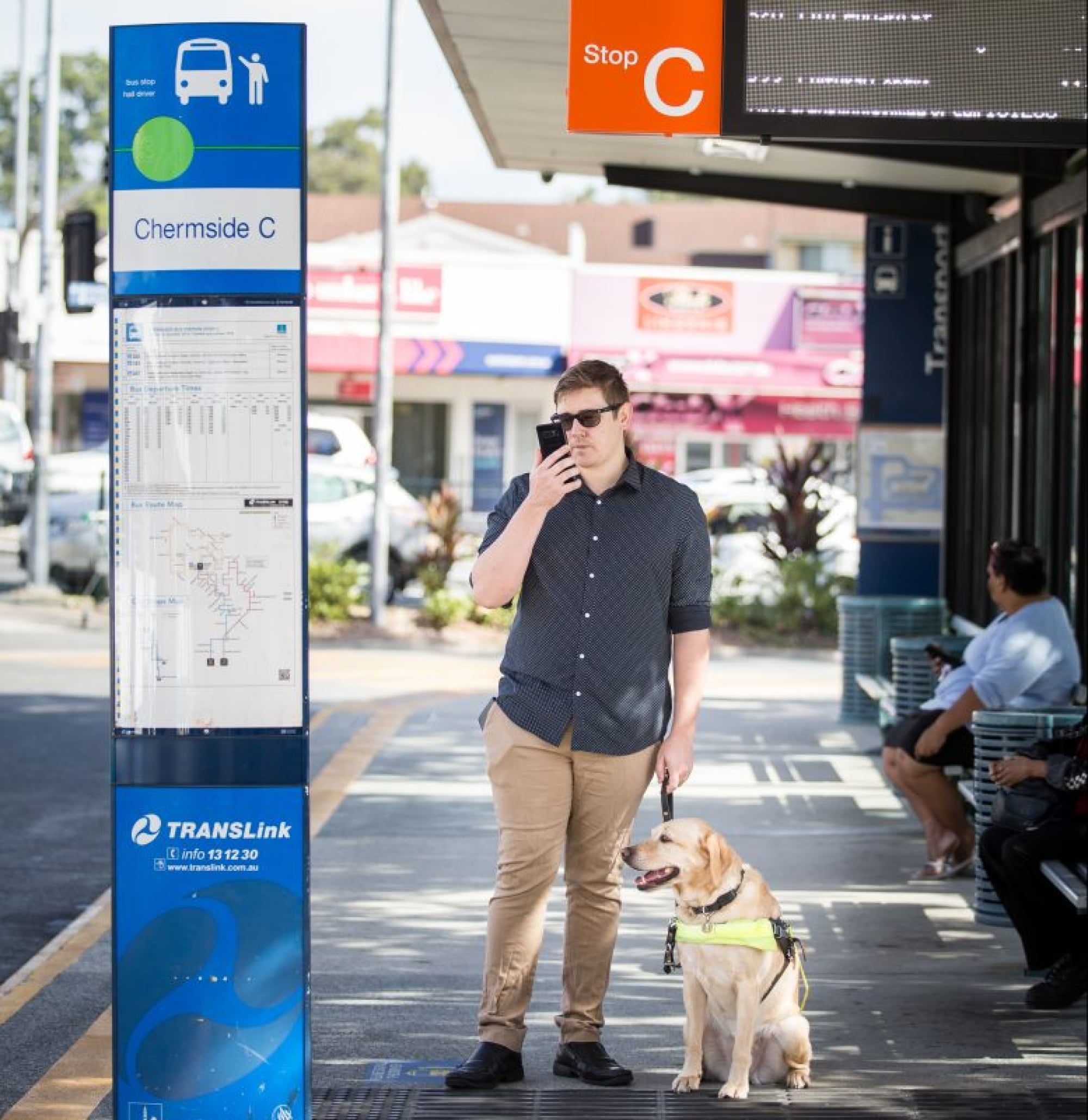 Person standing on a sidewalk with their yellow Guide Dog in harness who is seated next to them. The person is using their iPhone.