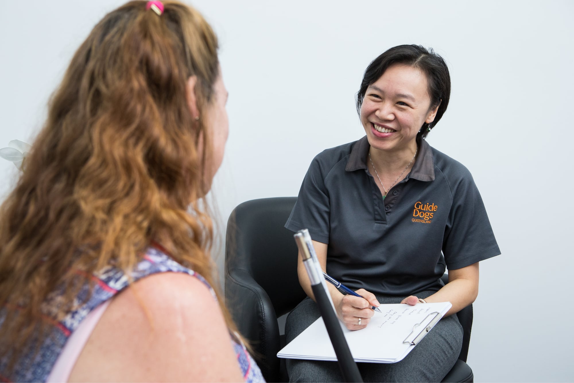 A person sitting with a Guide Dog's staff member. The staff member is smiling while the other person is not facing the camera.