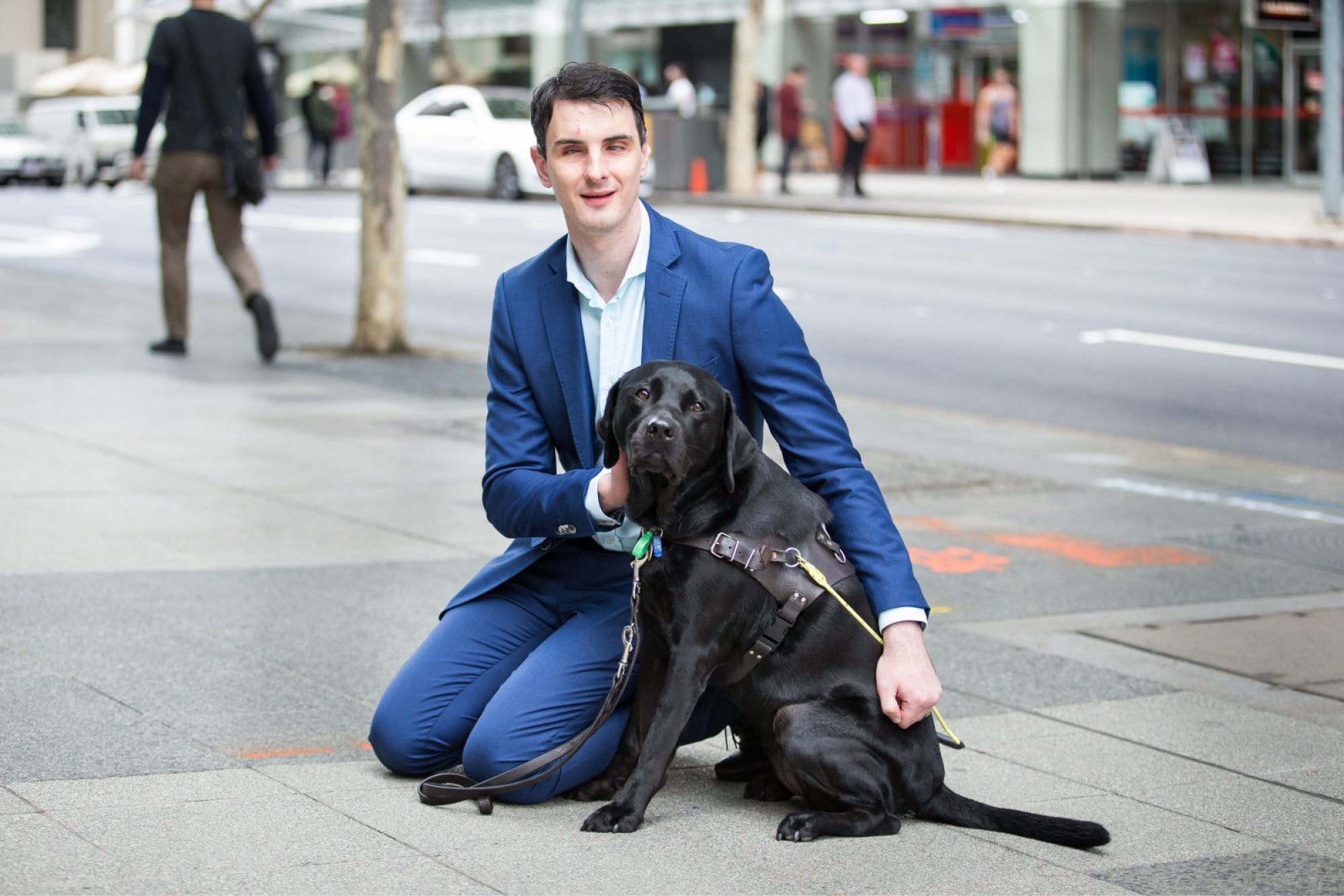 Person kneeling down outside next to a black Guide Dog in harness. The person and Guide Dog are looking at the camera.