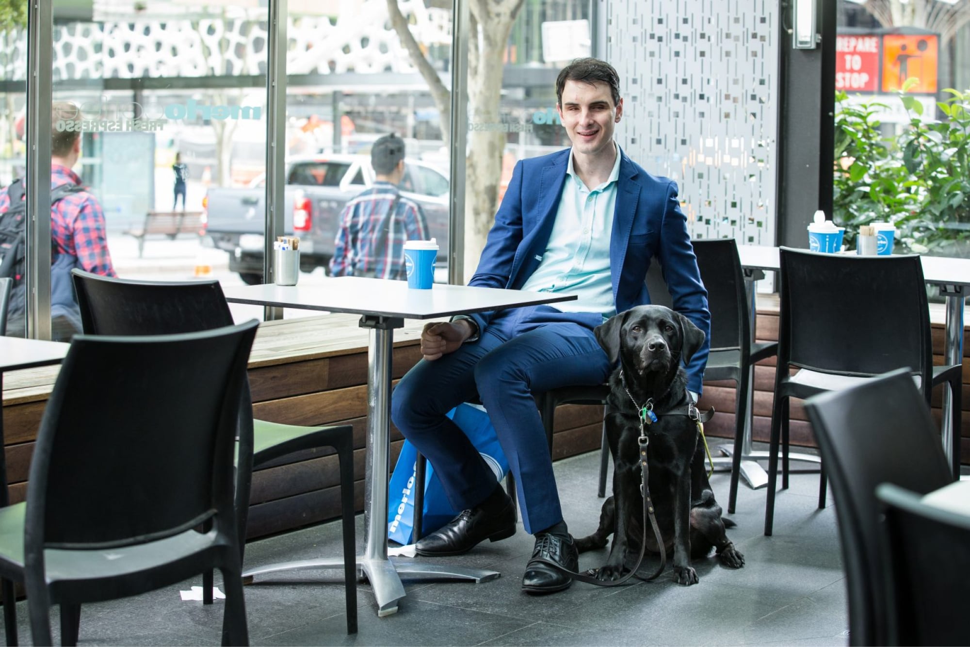 Person sitting at a table in a cafe with a black Guide Dog in harness by their side.