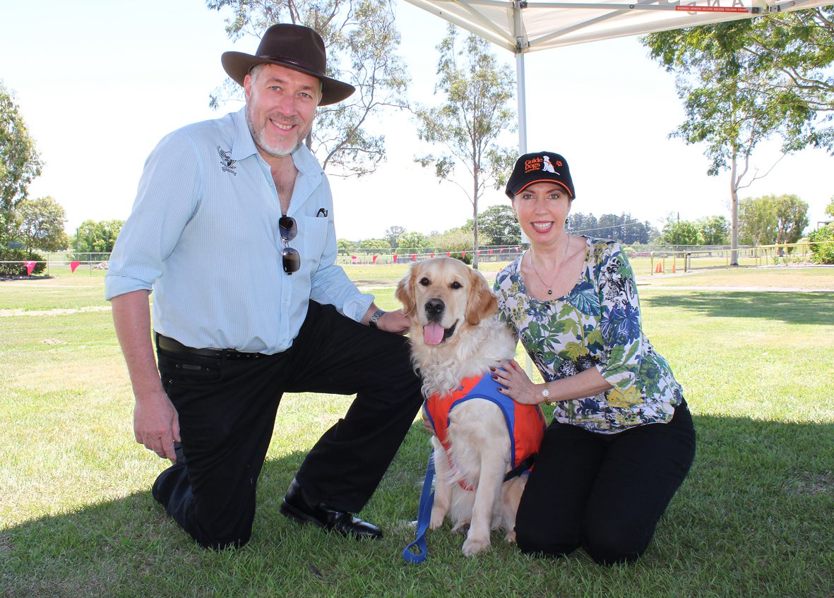 Image of a man and woman crouching on the grass beside a yellow Golden Retriever in a coat