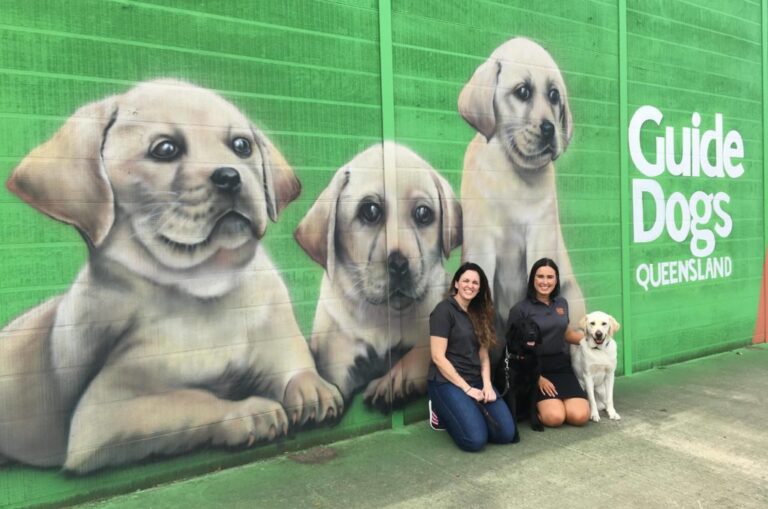 Image of two women and a yellow dog sitting in front of a colourful mural