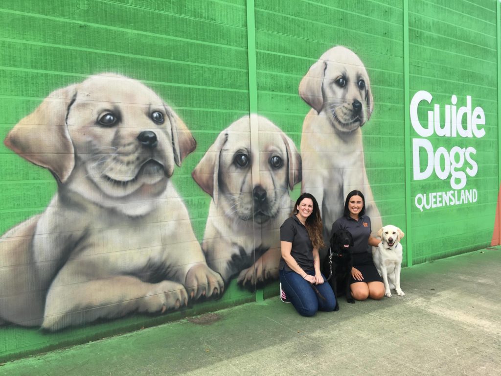 Image of two women and a yellow dog sitting in front of a colourful mural