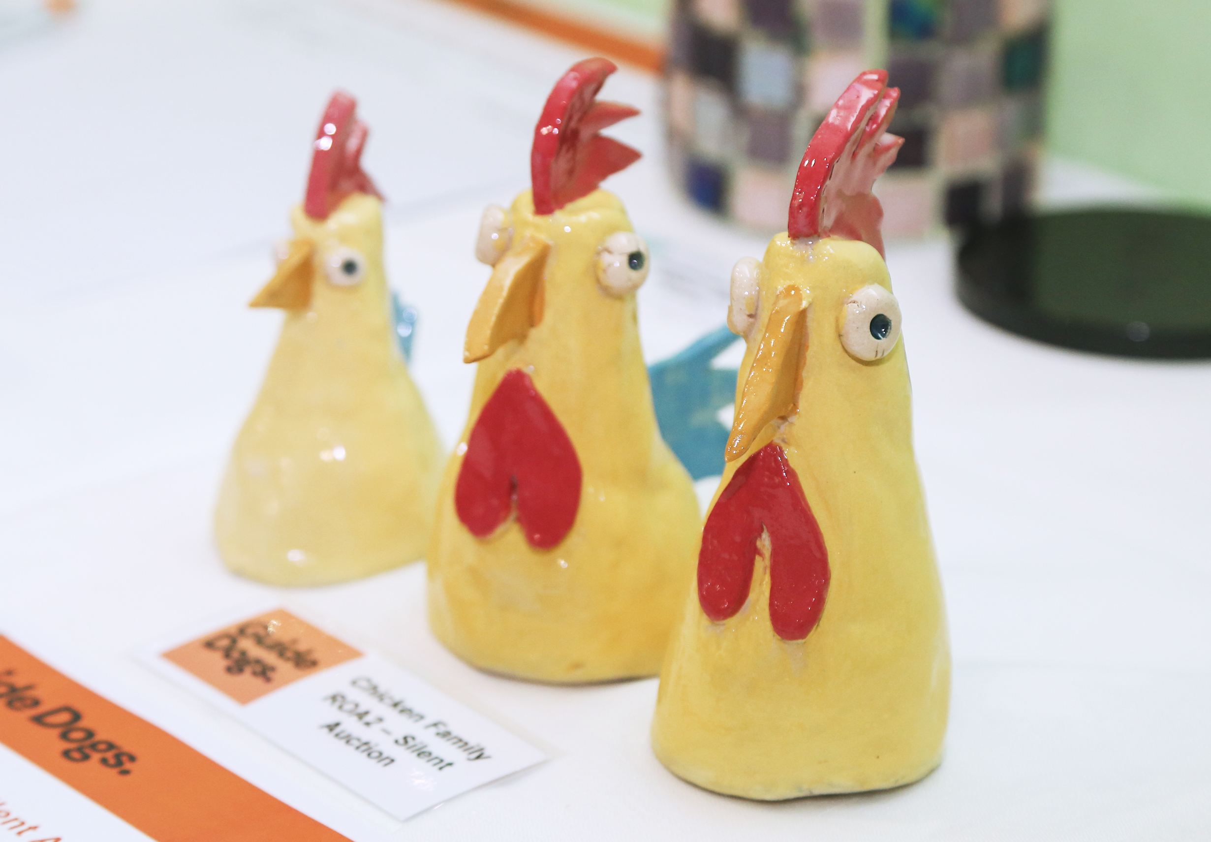 Three small, handmade ceramic chicken figurines standing in a row on a white surface. The yellow, cone-shaped chickens feature bright red combs and wattles, wide, cartoonish eyes, and small orange beaks. A small printed sign next to them reads 