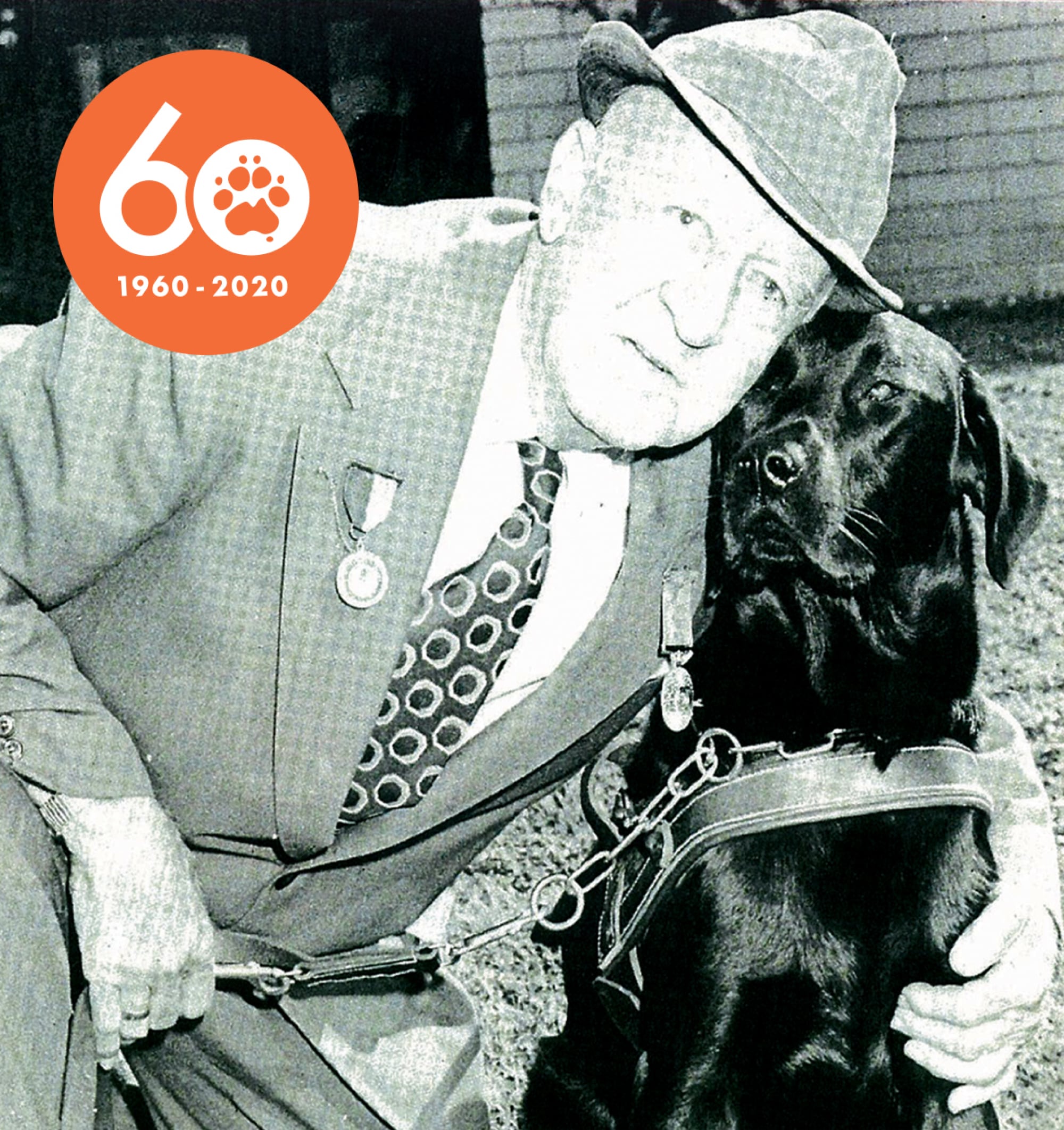 A 1960's black and white image of an older adult hugging a black labrador Guide Dog who is in a harness. The adult and dog are looking at the camera.