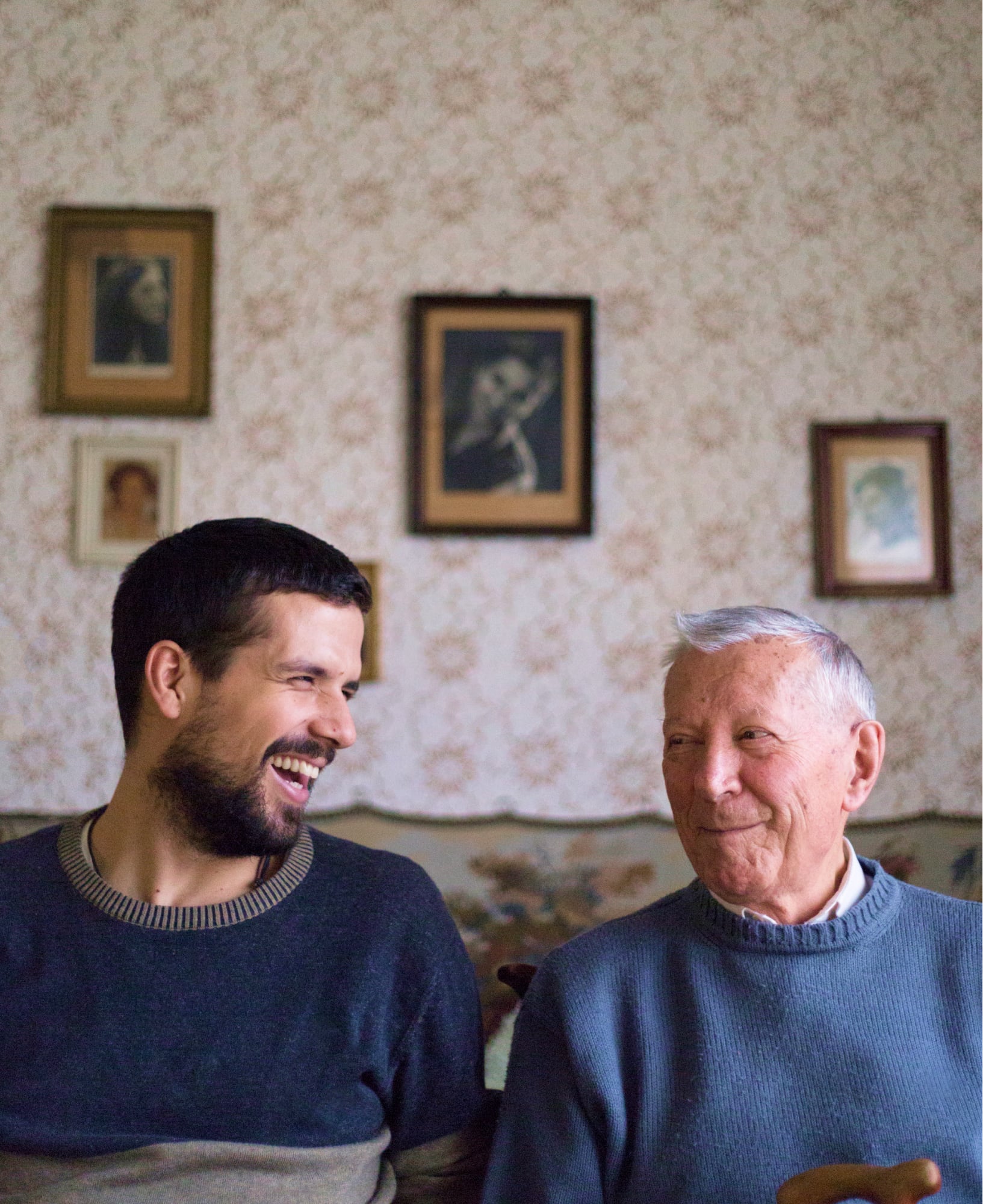 Two people, one adult & one older person, sitting on a couch looking at each other laughing.