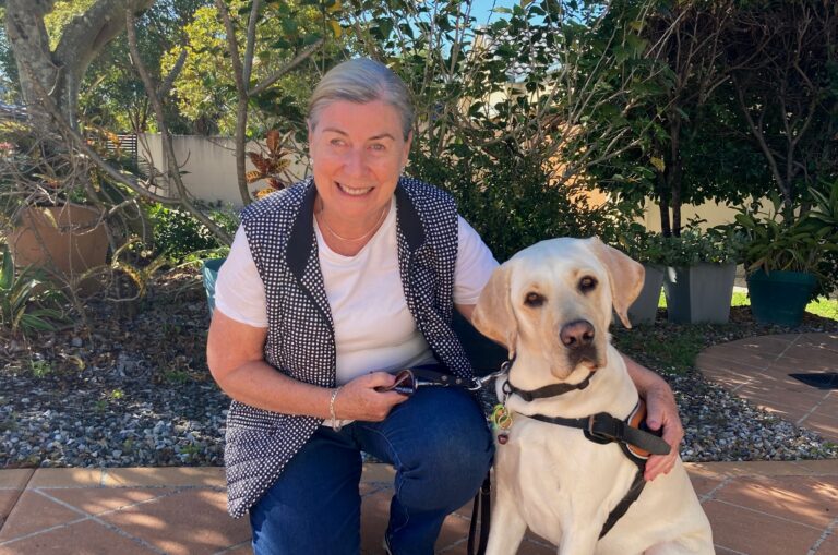 Anne-Marie is crouching next to Jerry, a yellow Guide Dog in front of a garden