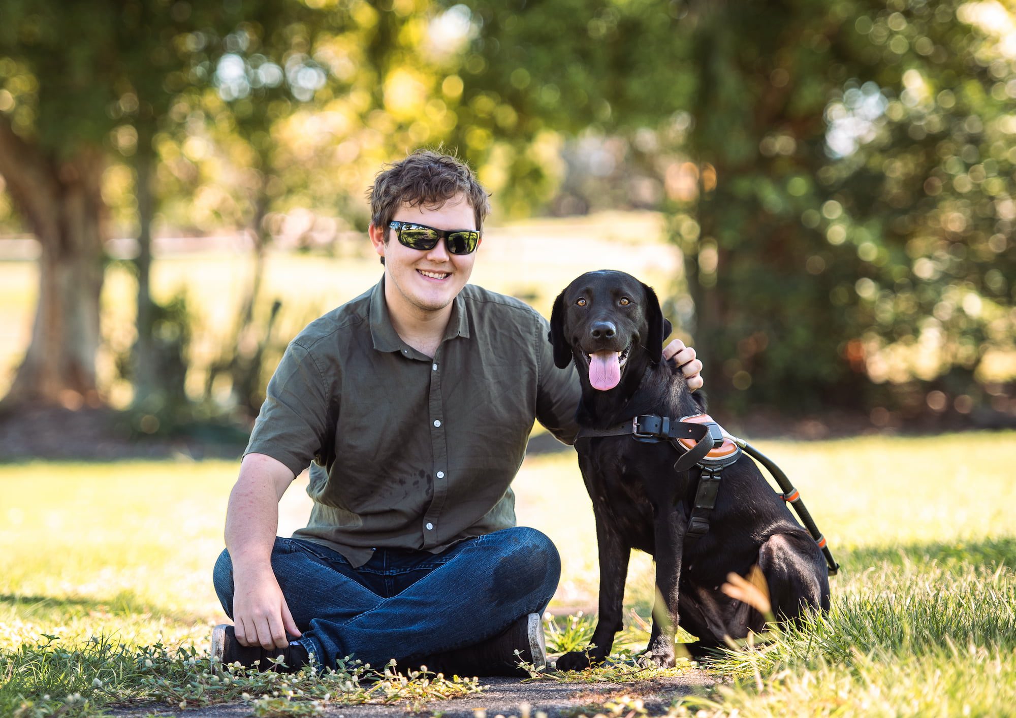 Brent and Guide Dog Jaycee sitting on the grass, looking at the camera, smiling.
