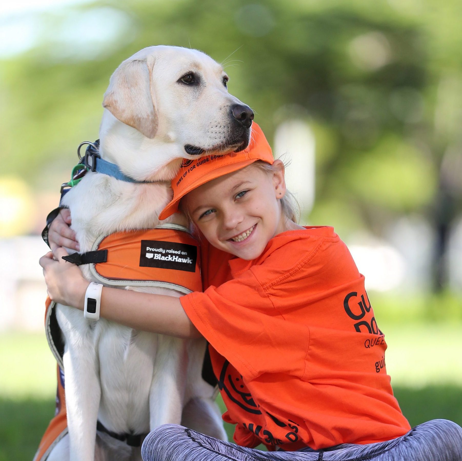 A smiling young girl wearing an orange t-shirt and cap sits on the grass, warmly hugging a white Labrador guide dog wearing an orange vest.