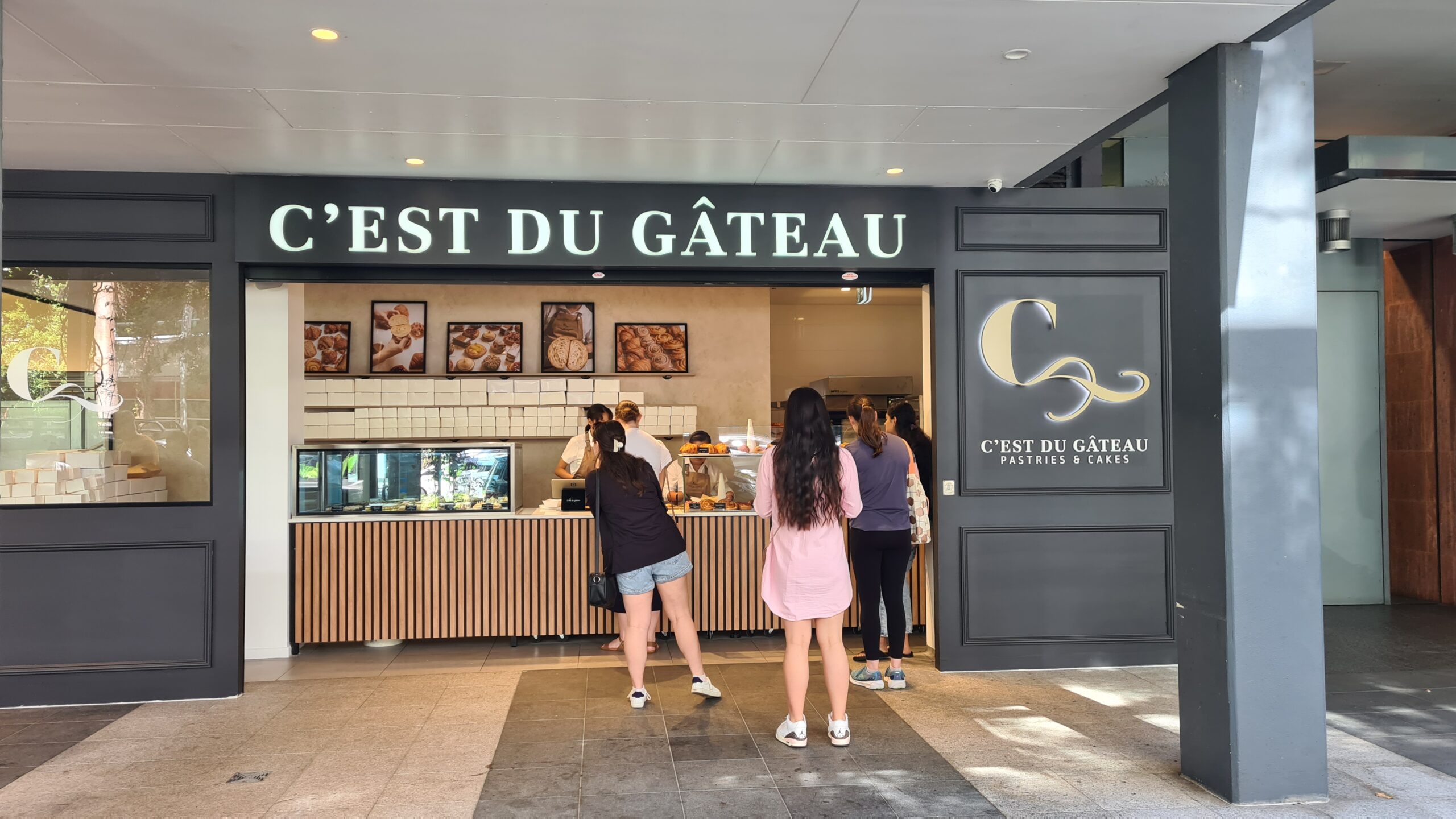 A street-level view of C'est Du Gateau bakery. The shop has a wide, step-free entrance and a long service counter. Several customers stand on the spacious, even-tiled pavement, highlighting the accessible and open floor plan.