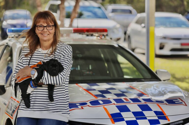 A woman holding Davey, a black Labrador puppy, in her arms. She is standing in front of a police car.