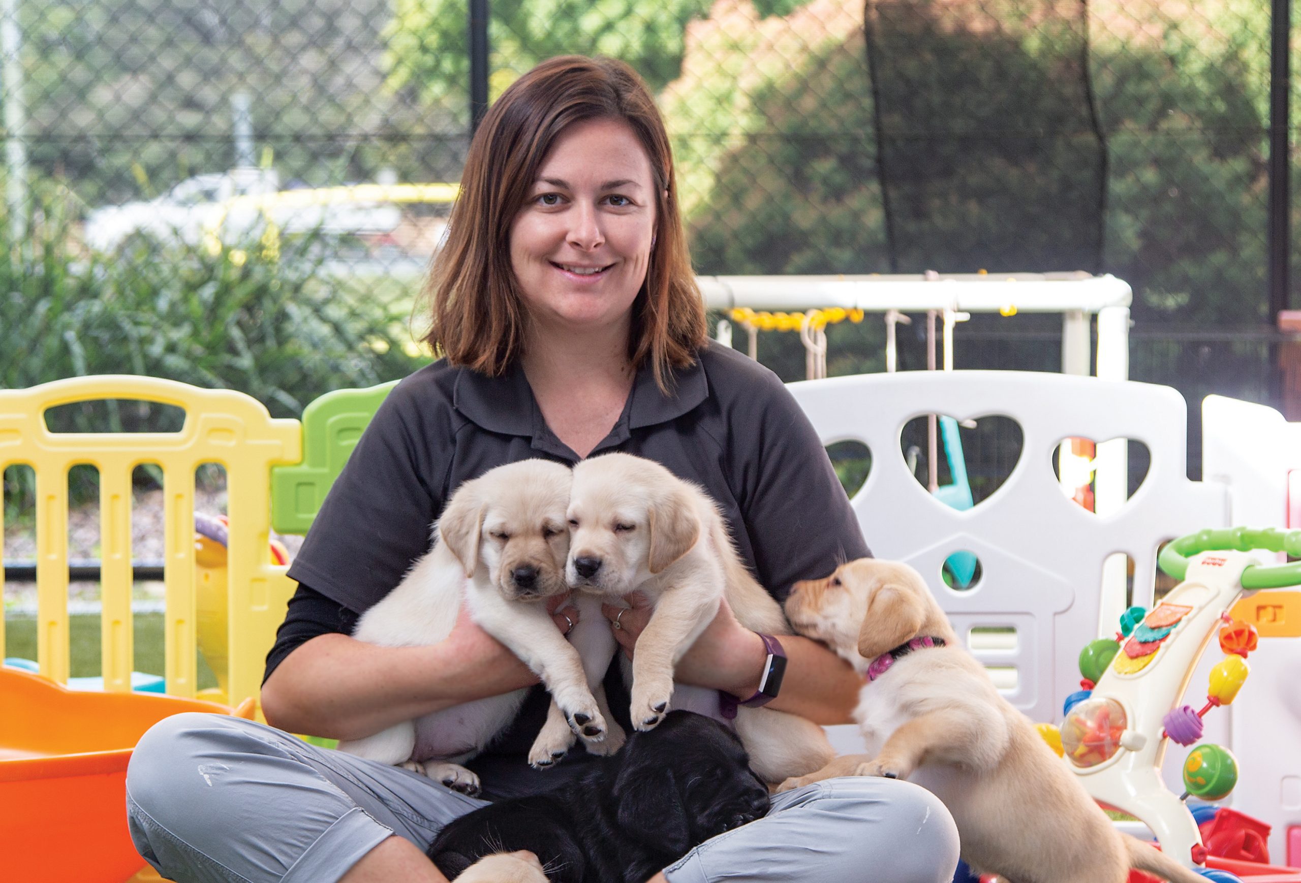 Image of Fiona holding two puppies in the S litter with another beside her, sitting the puppy play area.