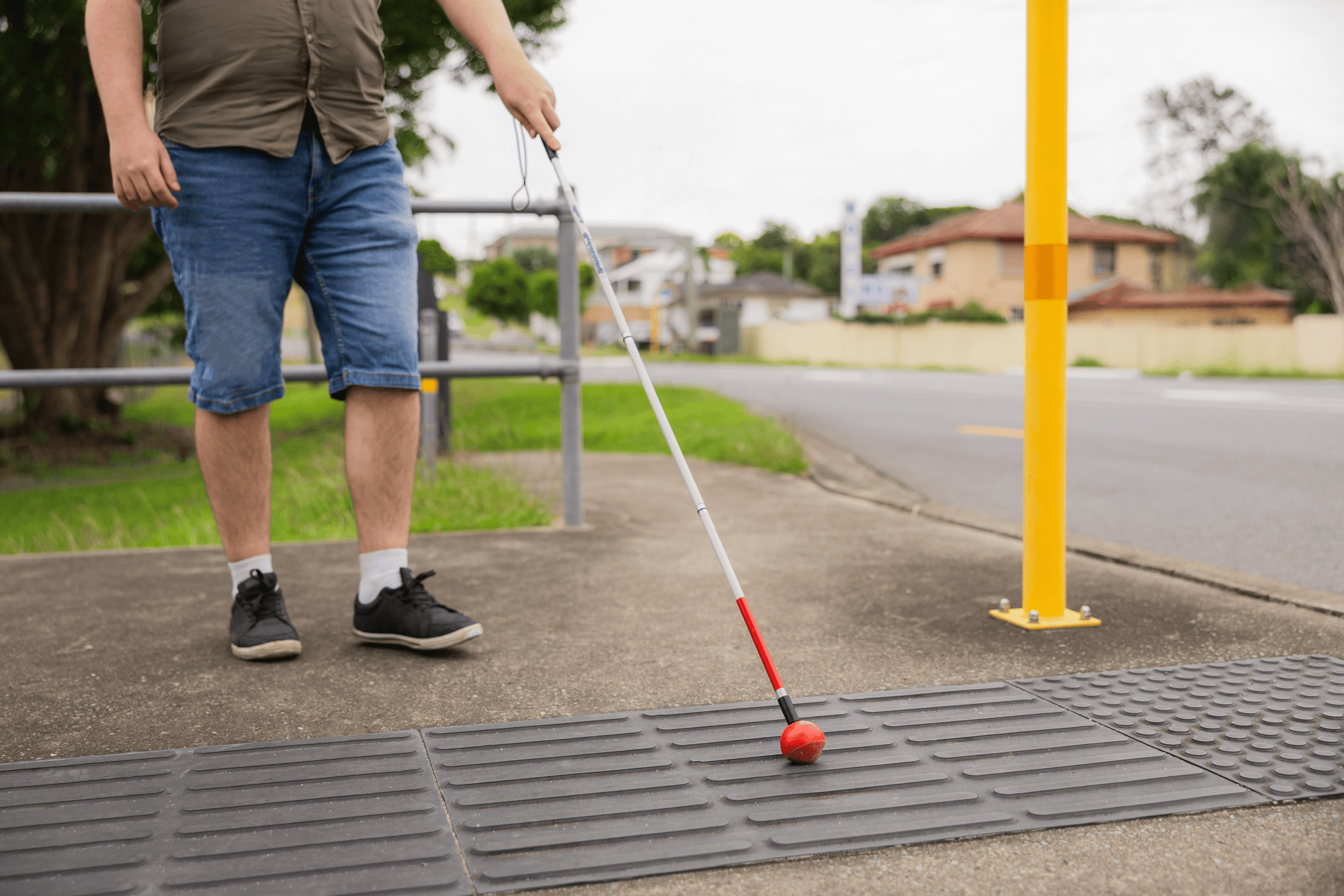 Image shows a person using a white cane with a red tip to navigate tactile paving at a street crossing, indicating an accessible pedestrian path.