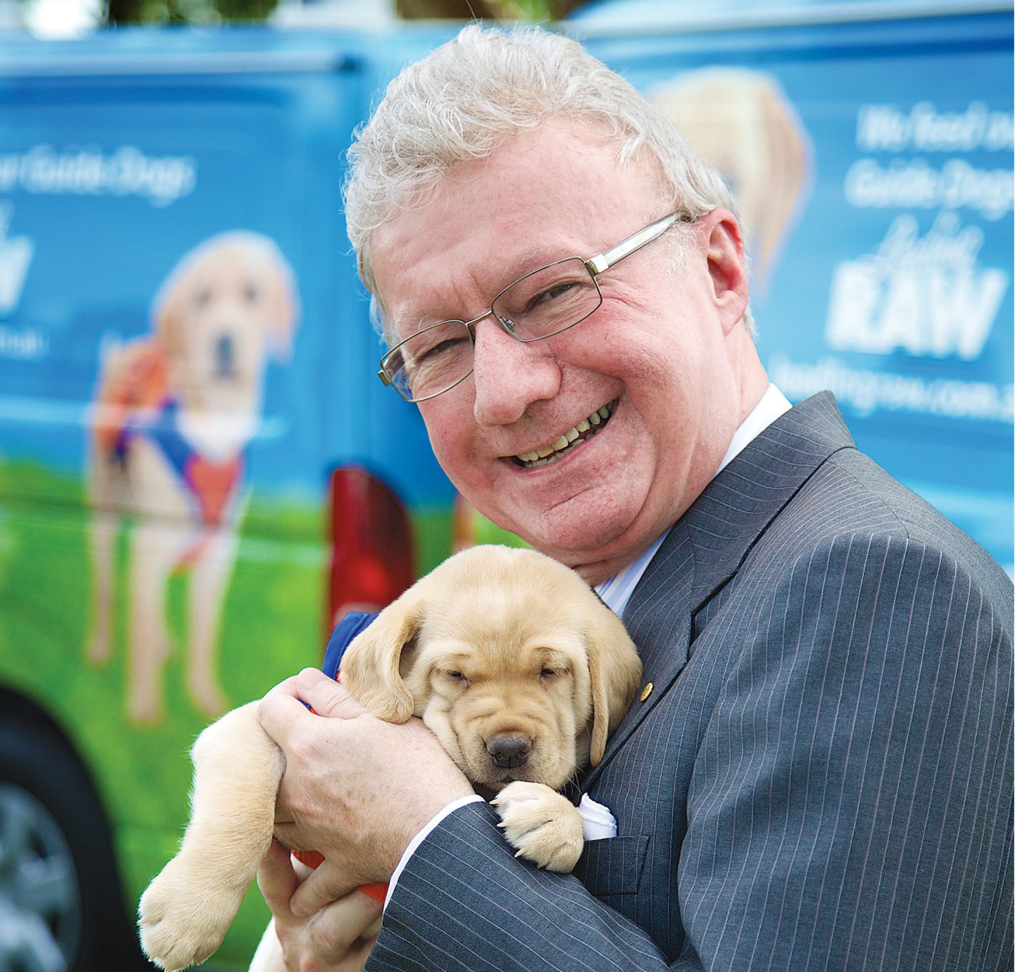 Governor of Queensland, Paul de Jersey AC, holding an eight week old yellow labrador puppy. His Excellency is smiling while looking at the camera.