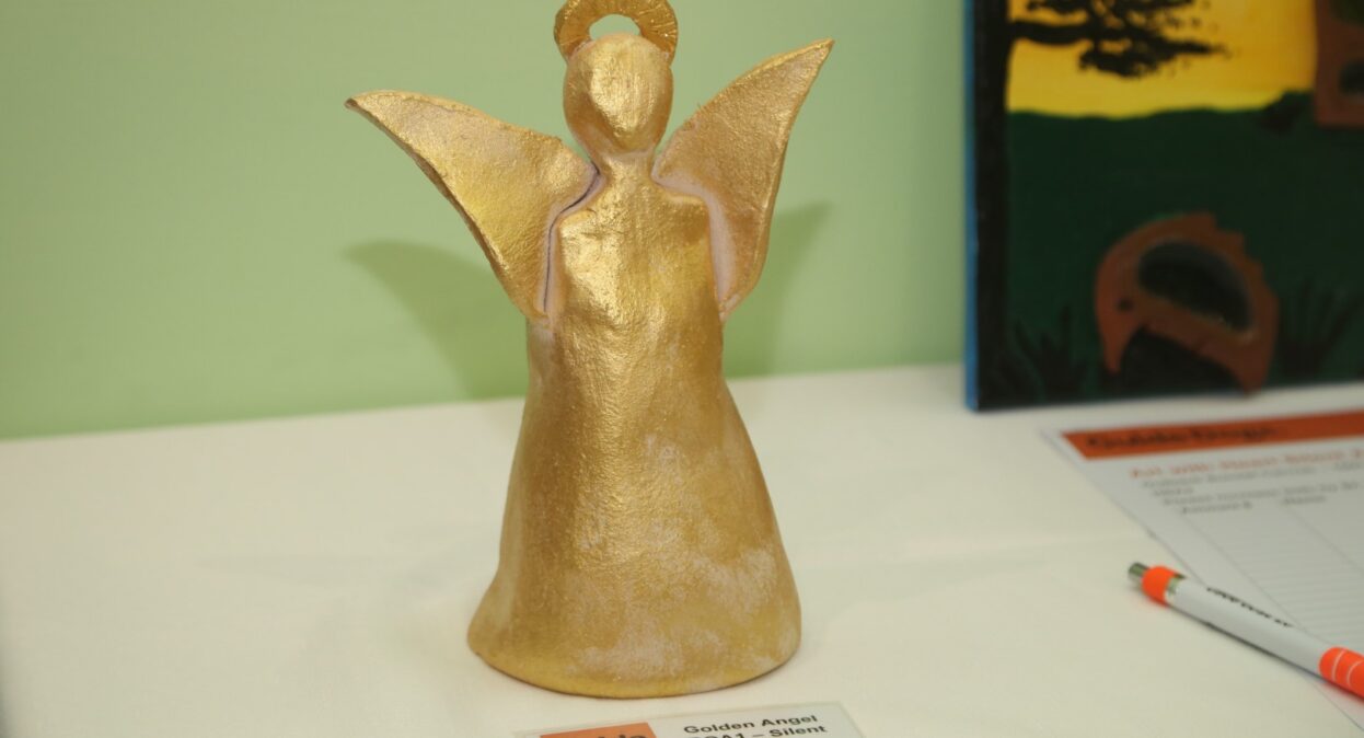 A small, handmade golden ceramic angel figurine with a halo stands on a white tablecloth next to a "Guide Dogs" silent auction label.