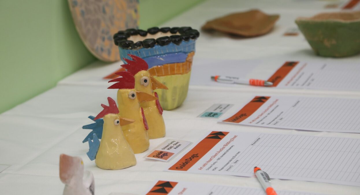 A row of three handmade ceramic chicken figurines sits on a table alongside silent auction bidding sheets and pens.