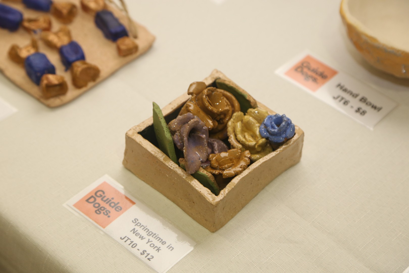 A small square ceramic box filled with handmade glazed pottery roses in brown, purple, yellow, and blue rests on a tablecloth next to its display label.