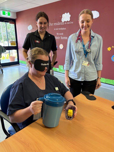 A staff member wearing a blindfold practices handling a jug while two colleagues observe.