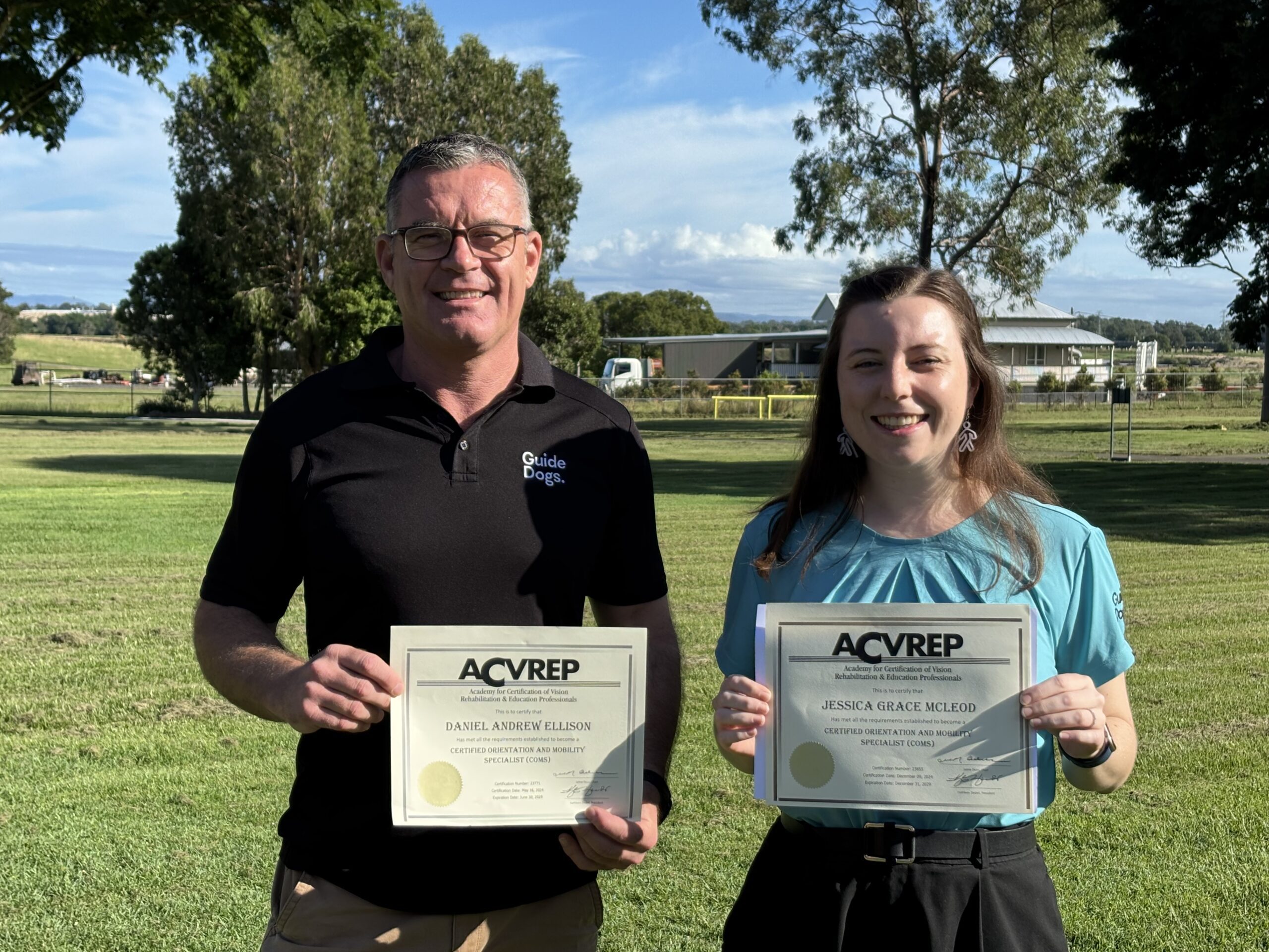 Daniel Ellison and Jessica McLeod, wearing Guide Dogs uniforms, hold ACVREP COMS certificates after achieving certification as Orientation and Mobility Specialists.
