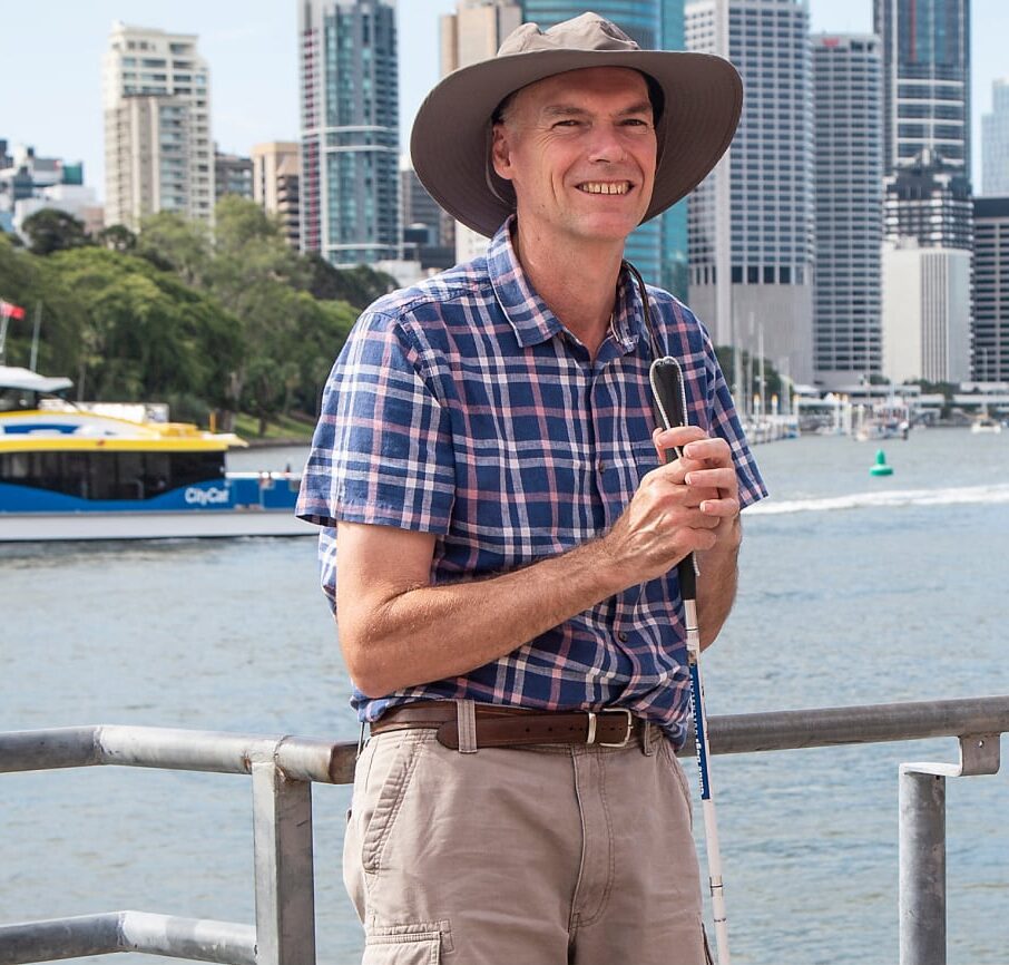 A Client stands outside holding their cane to their chest as they smile to the camera. A scenery or Brisbane River and skyline is behind them.