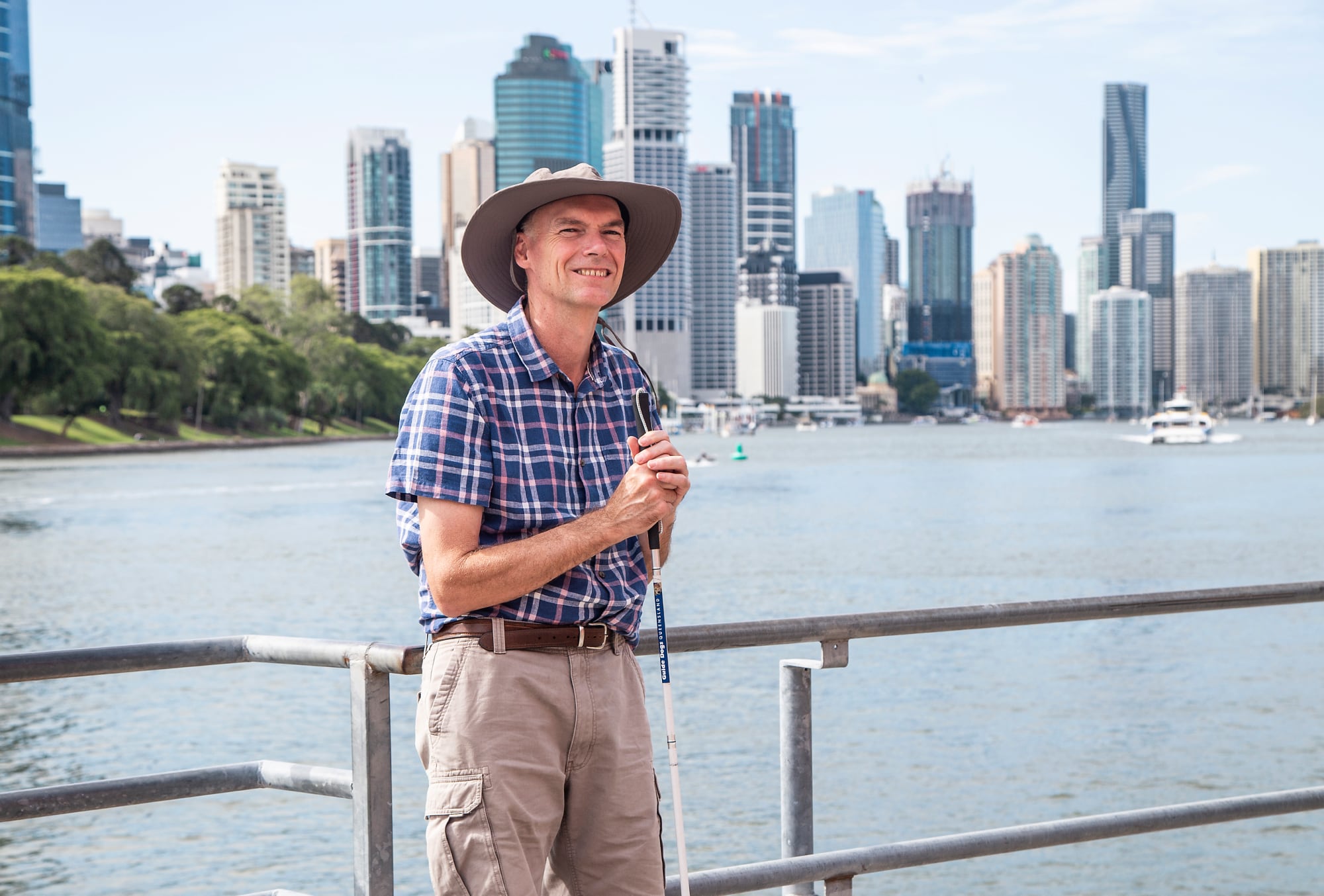 A Client stands outside holding their cane to their chest. A scenery or Brisbane River and skyline is behind them.
