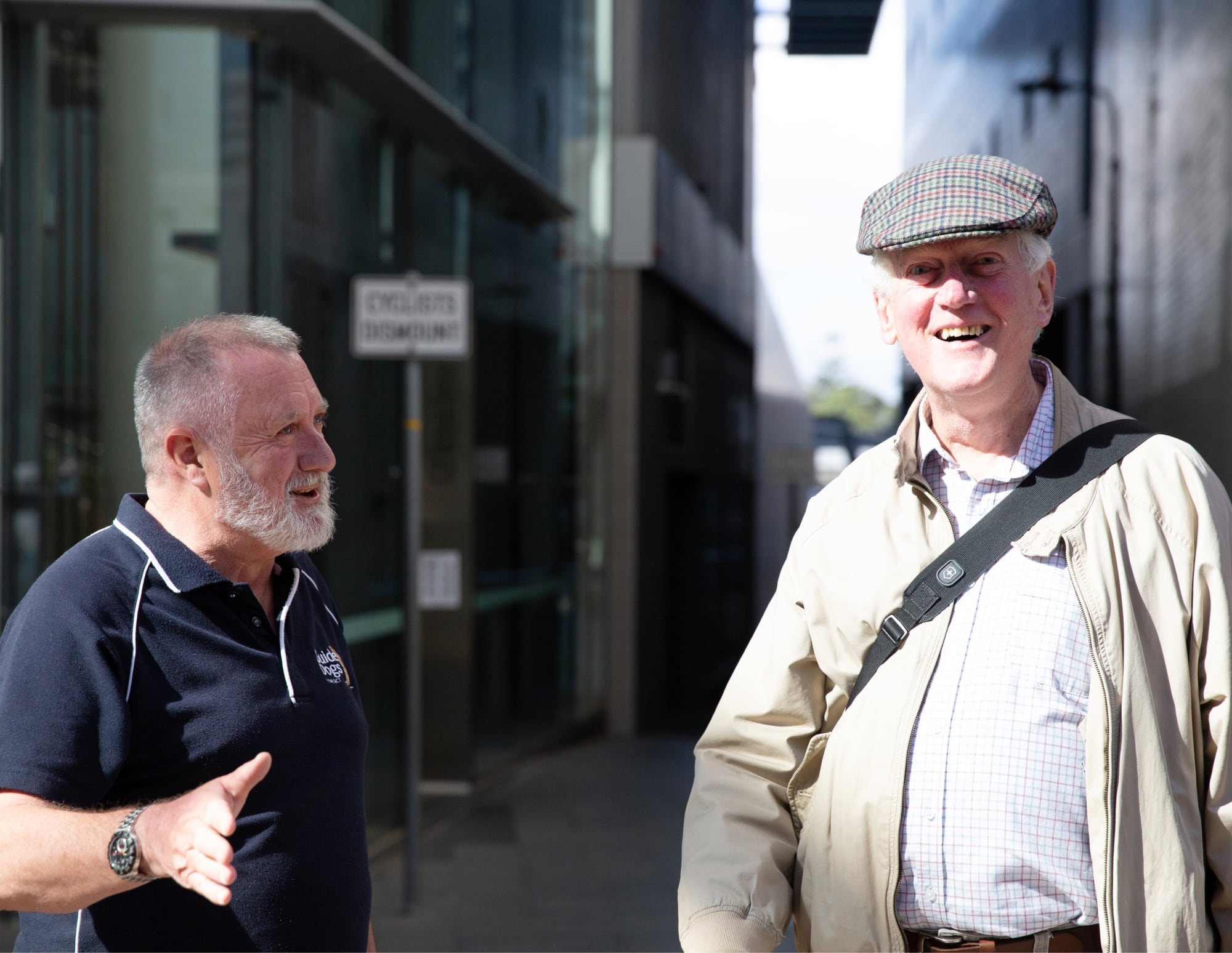 A Guide Dogs staff members is talking to an older adult in the street. The an older adult is looking at the camera with a smile on his face.