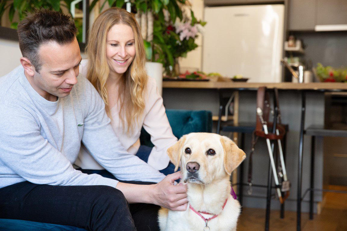 Image shows a man and woman gently petting a yellow Labrador in a kitchen-living space, with a guide dog harness visible nearby.