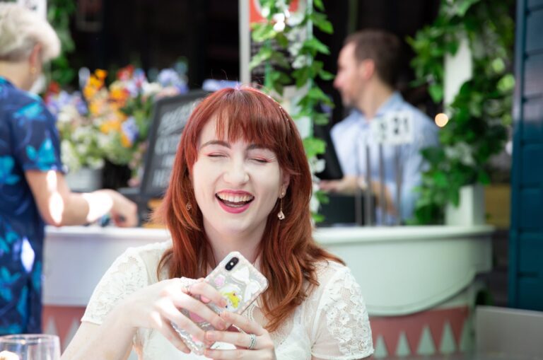 An adult sitting outside at a cafe laughing while holding their mobile phone.
