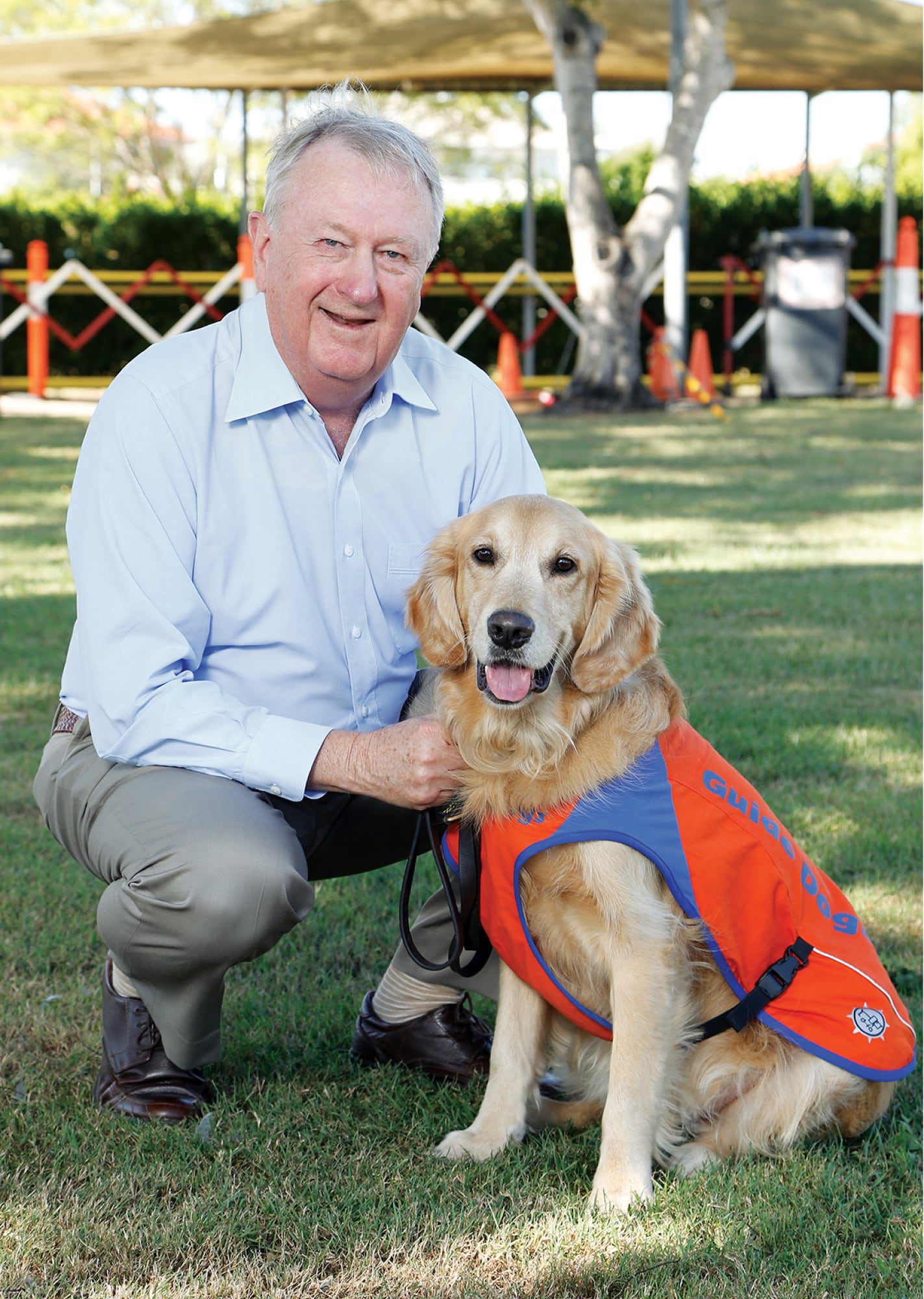 Guide Dogs Queensland Board President, Richard Anderson OAM, kneeling outside next to a yellow labrador Guide Dog in Training. The dog is wearing a Guide Dog in Training jacket. Both the dog and Richard are looking at the camera and Richard is smiling.
