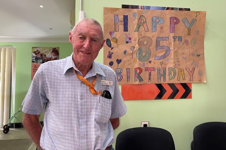 Guide Dogs Client, Ray is standing in front of a handmade sign which reads 'Happy 85th Birthday'.