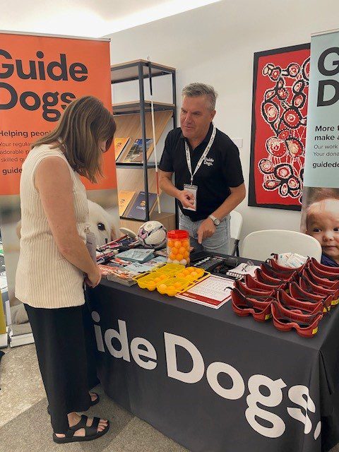 Two people standing at a Guide Dogs stall. The woman is looking at products on the table.