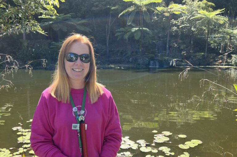 Sarah is standing in front of a pond surrounded by gardens and small waterfalls. Sarah is holding her White Cane and smiling at the camera