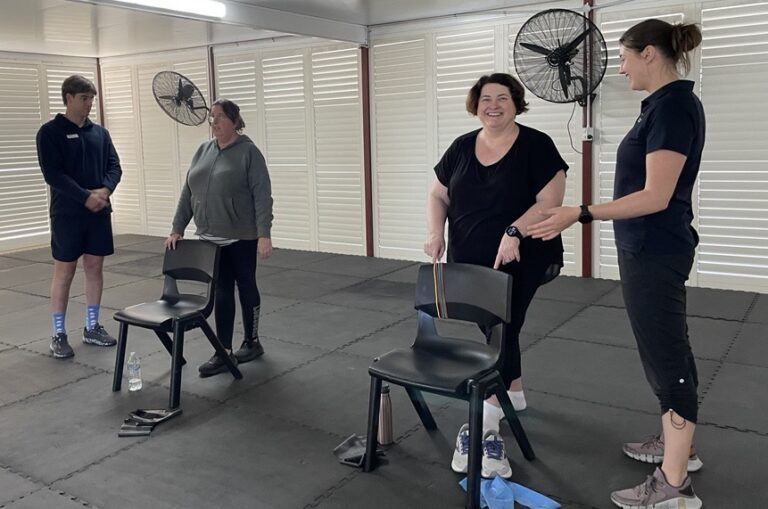 Two clients are pictured standing behind black chairs, each with an exercise physiologist next to them.