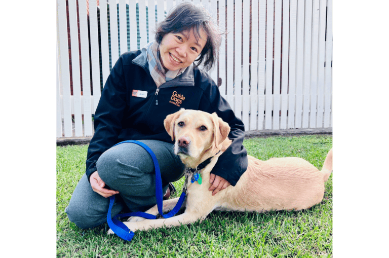 Clinical Psychologist, Dr. Sau Kuan Cheong (Janet) is sitting on the grass in front of a white fence. Jasmine, a yellow Labrador, lies by Janet's side.