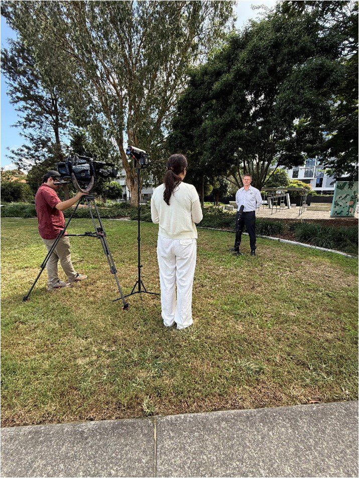 Image shows a news crew filming an outdoor interview in a park, with a woman facing a man speaking on camera as a videographer records the scene.