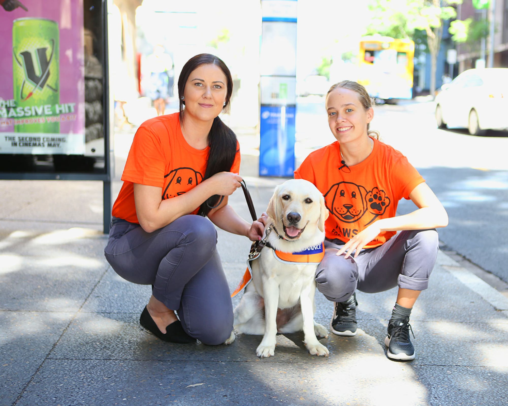 Two people kneeling next to a yellow Guide Dog in training who is wearing a Guide Dog in Training jacket. They are outside on the footpath. The people are smiling while looking at the camera.