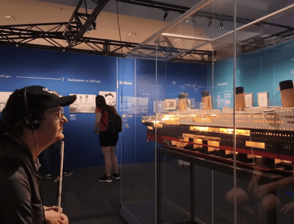 Man with a white cane and headset observing a detailed, illuminated model of the Titanic in a museum exhibit.