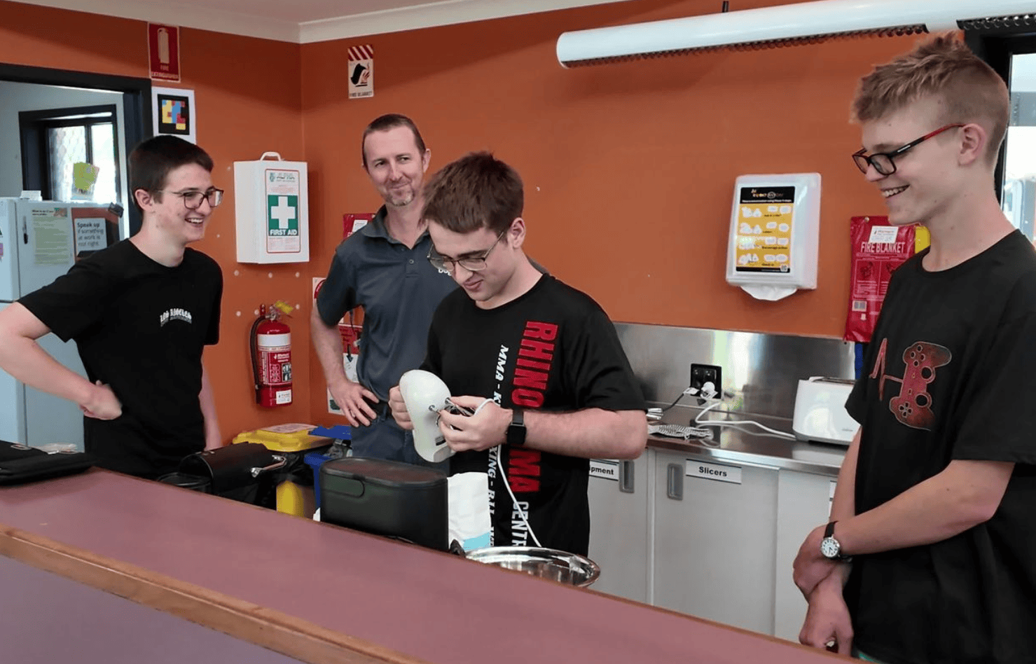 Teen boy examines an electric hand mixer while others and an instructor smile nearby in a kitchen classroom.