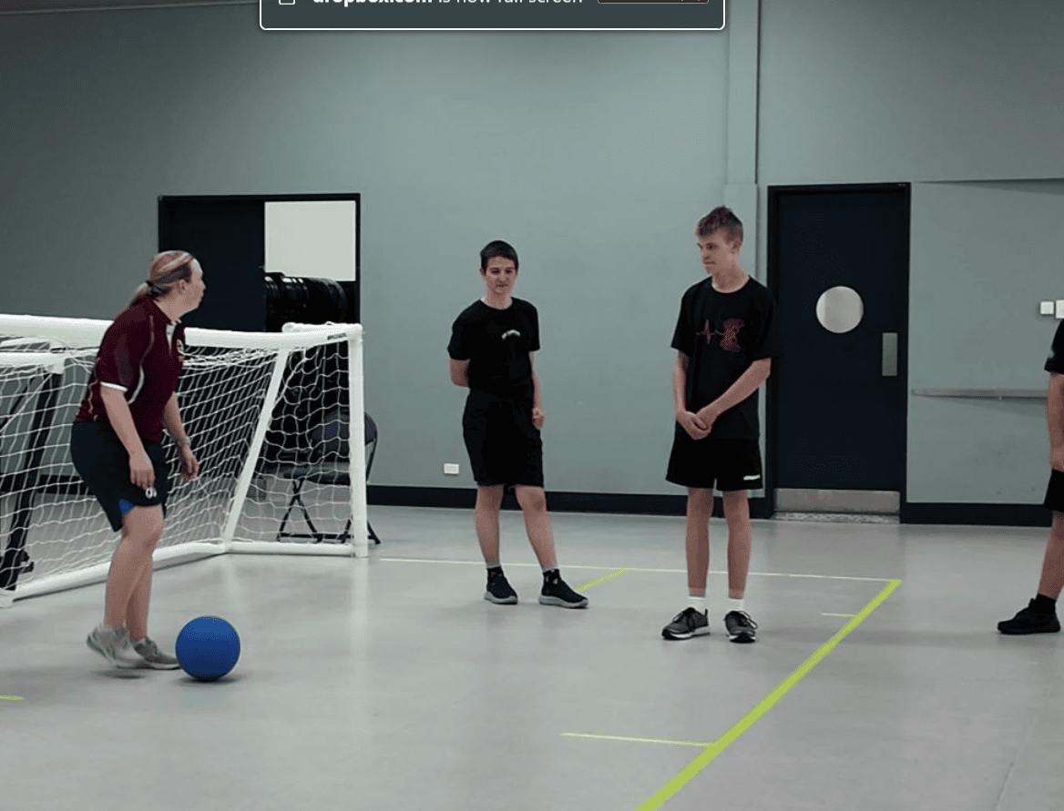 Students and an instructor play goalball in a gym, with one player preparing to roll a blue ball toward the others.