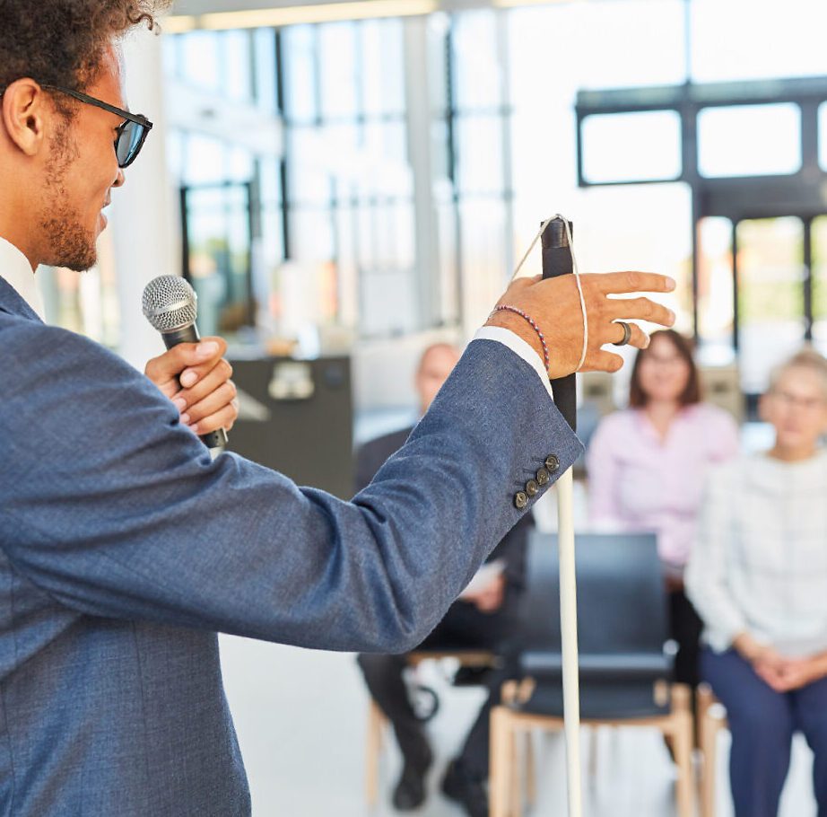Man with a cane address an audience sitting in front of him.